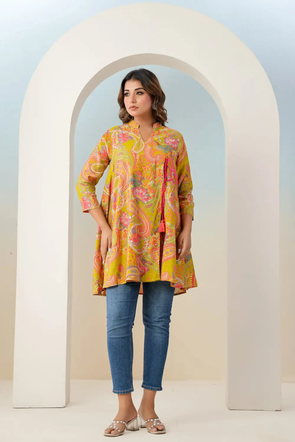 Woman wearing a colorful floral dress and jeans standing in front of a white archway.